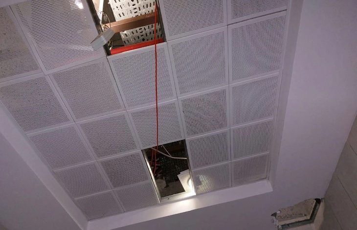 Fotoğrafta, tavanı oluşturan kare formlu, delikli panellerden biri yerinden çıkmış ve tavandaki kablolar ile yapı elemanları açık şekilde görülüyor; ayrıca bir aydınlatma armatürü de boşluktan aşağı doğru ışık veriyor.