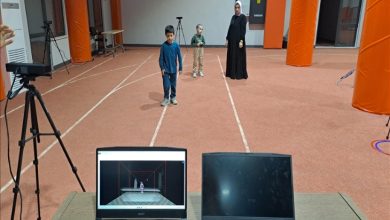 Geniş bir iç mekânda atletizm pistlerini andıran çizgiler var. Ortada, farklı yaşlarda iki çocuk ve arkalarında siyah uzun elbiseli bir yetişkin kadın ayakta duruyor. Ön planda bir masa üzerinde iki dizüstü bilgisayar bulunuyor. Bilgisayarlardan birinin ekranında, hareket yakalama/simülasyon benzeri bir görüntüyü gösteren bir sahne açık.