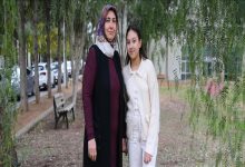 Ağaçların altında, park benzeri bir alanda yan yana duran iki kişi görülüyor. Biri başörtülü yetişkin bir kadın, diğeri açık renk kıyafetli genç bir kız; kameraya dönük, sakin ve vakur bir şekilde poz veriyorlar. Arka planda ağaçlar, bank ve park halindeki araçlar yer alıyor.