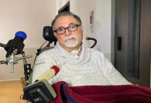 ALS hastalığıyla mücadele eden emekli göz doktoru Alper Kaya, tekerlekli sandalyede oturuyor, boynunda tıbbi bir solunum aparatı bulunuyor ve üzerinde açık renkli bir kazak var.