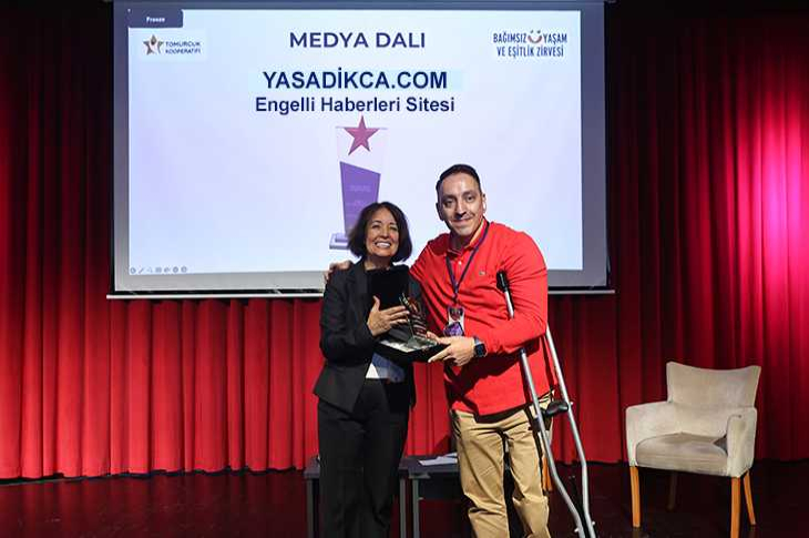 Bir sahnede, ödül töreninde iki kişi birlikte plaket tutup gülümsüyor; arkada perdede “yasadikca.com Engelli Haberleri Sitesi” yazısı ve kırmızı fon perdeler görülüyor.