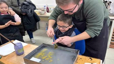 Bir atölye sınıfında, bir eğitmen küçük bir çocuğun elini yönlendirerek sulu boya çalışması yapmasına yardımcı oluyor. Çocuk gözlük takıyor ve gülümseyerek fırçayı tutuyor. Masada boya kapları ve tepsi içinde su yüzeyine yayılan renkli desenler var. Arkada diğer çocuklar ve veliler izliyor.