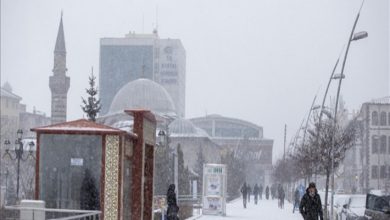 Yoğun kar yağışı altında bir şehir meydanı görülüyor. Minareli bir cami, kubbeli yapılar ve modern binalar arka planda siluet gibi. Yoldan yürüyen birkaç kişi kalın montlarla karın içinde ilerliyor. Hava gri, sessiz ve ağır.