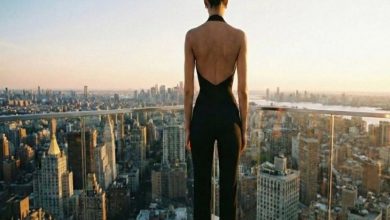 Yüksek bir binanın terasında, sırtı kameraya dönük bir kadın şehir manzarasına bakıyor. Siyah, sırtı açık bir kıyafet giymiş; önünde gün batımında uzanan yoğun gökdelen silueti ve cam korkuluklar görülüyor.