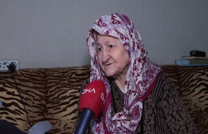 Evinde röportaj veren yaşlı bir kadın, başörtüsüyle koltukta otururken DHA mikrofonuna konuşuyor; yüzündeki ifade yaşadığı zorluğu ve yardım beklentisini yansıtıyor.