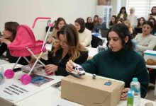 Sınıf ortamında, sıralara dizilmiş öğrenciler dikkatle kendi geliştirdikleri maket projelerle ilgileniyor. Ön planda iki genç kadın; biri pembe oyuncak bebek arabasıyla, diğeri karton bir platform üstünde küçük bir ambulans modeliyle çalışıyor. Masada su şişeleri, notlar ve proje kartları var.