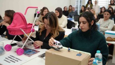 Sınıf ortamında, sıralara dizilmiş öğrenciler dikkatle kendi geliştirdikleri maket projelerle ilgileniyor. Ön planda iki genç kadın; biri pembe oyuncak bebek arabasıyla, diğeri karton bir platform üstünde küçük bir ambulans modeliyle çalışıyor. Masada su şişeleri, notlar ve proje kartları var.