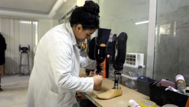 Beyaz önlük giymiş bir Sena Kanalıcı, atölye ortamında bir çalışma tezgâhı üzerinde protez bacaklar üzerinde dikkatle çalışmaktadır.