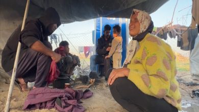 Geçici bir çadırın altında, yerde ateş üzerinde yemek hazırlanırken bir kadın gülümseyerek oturuyor, yanında bir adam küçük bir çocuğu kucağında tutuyor ve diğerleri günlük işlerle meşgul oluyor.