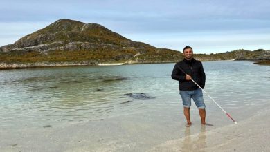 Görme engelli öğretim üyesi Dr. Önder İşlek, sahilde, berrak sığ suların içinde çıplak ayakla dururken, elindeki beyaz bastonla poz veriyor.