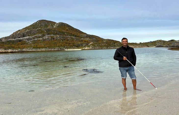 Görme engelli öğretim üyesi Dr. Önder İşlek, sahilde, berrak sığ suların içinde çıplak ayakla dururken, elindeki beyaz bastonla poz veriyor.