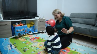 Bir oturma odasında, renkli oyun matı üzerinde diz çöken bir yetişkin, karşısında oturan küçük bir çocukla oyun oynuyor; etrafta oyuncaklar, arkada televizyon ve koltuk var.