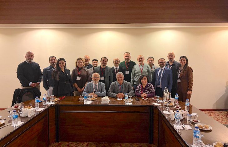 Toplantı salonunda, uzun bir masa etrafında toplanmış kalabalık bir grup görülüyor. Ön sırada oturanlar, arkada ayakta duranlarla birlikte kameraya poz vermiş. Masa üzerinde su şişeleri ve ikramlar var; ortam resmi ve kurumsal bir toplantı havasında.