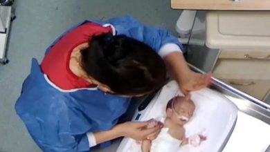 Üstten çekilmiş görüntüde, hastane ortamında bir sağlık çalışanının yenidoğan bir bebeğe bakım uyguladığı görülüyor; bebek kuvöz benzeri bir yatakta, çalışanın elleri bebeğin baş ve kol bölgesinde.