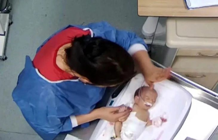 Üstten çekilmiş görüntüde, hastane ortamında bir sağlık çalışanının yenidoğan bir bebeğe bakım uyguladığı görülüyor; bebek kuvöz benzeri bir yatakta, çalışanın elleri bebeğin baş ve kol bölgesinde.