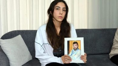 Gri bir koltukta oturan genç bir kadın, yüzünde ağır bir hüzünle, ellerinde çerçeveli bir erkek fotoğrafı tutuyor. Sade bir oda, sessiz bir yas; bakışları kayba kilitlenmiş, zaman durmuş gibi.