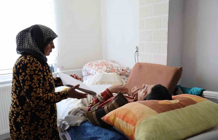 Yaşlı babaanne Zeynep Atmaca, aydınlık bir odada yatağa uzanmış torununa elindeki kâğıtlarla ilgilenirken şefkatle yanında durmaktadır.