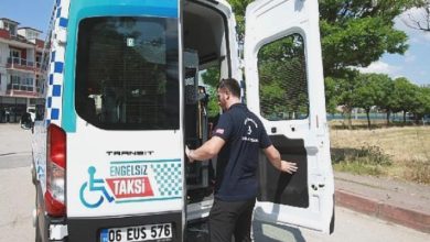 Bir minibüs tipi “Engelsiz Taksi”nin arka kapısı açık; görevli araçtan inip binmeye yardımcı oluyor. Belediye logosu ve tekerlekli sandalye simgesiyle, erişilebilir ulaşım hizmeti vurgulanıyor.