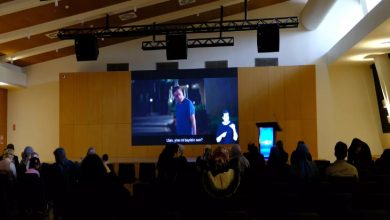 Büyük bir salonda toplanan izleyiciler, sahnedeki dev ekranda betimlemeli film gösterimini takip ediyor; loş ortamda erişilebilirliğe odaklanan bir etkinlik havası var.