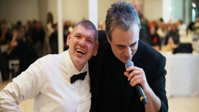 Sahne ışıkları altında, iki adam yan yana. Biri beyaz gömlek ve papyonla geniş bir gülümsemeyle kalabalığa bakıyor; diğeri siyah kıyafetle mikrofonu elinde, eğilerek şarkı söylüyor. Arkada bulanık bir salon dolusu insan, ön planda sıcak ve samimi bir an.