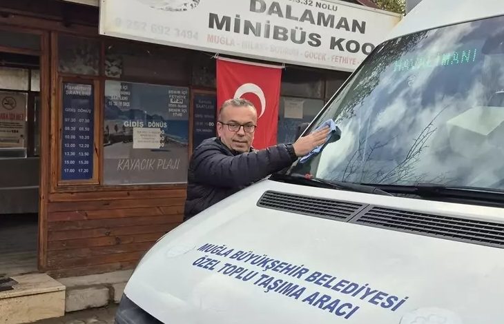 Minibüs durağı önünde bir şoför, belediyeye ait toplu taşıma aracının ön camını siliyor. Arkada Dalaman Minibüs Kooperatifi tabelası ve Türk bayrağı görünüyor.
