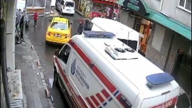 Dar bir sokakta güvenlik kamerasından çekilmiş görüntüde, yol kenarında park etmiş bir ambulansın önünde duran sarı bir taksi ve çevrede yürüyen birkaç yaya görülüyor. Otizmli Muhammed Ömer Karayel yol ortasında durarak trafik akışını sağlıyor