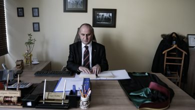 Bir ofiste masasının başında oturan takım elbiseli bir erkek, önündeki evrakları parmaklarıyla okuyup düzenliyor. Masada ofis eşyaları ve cübbe benzeri bir giysi dururken, arka planda siyah-beyaz fotoğraflar sade ve ciddi bir atmosfer yaratıyor.