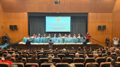 Büyük bir konferans salonu, sahnede uzun bir masa ve arkasında sıralanmış komisyon üyeleri. Karşılarında dolu bir salon; izleyiciler dikkat kesilmiş, kameralar ve telefonlar havada. Resmi, ciddi ve karar anına benzer bir atmosfer: konuşulanlar sadece söz değil, hayatlara dokunacak meseleler gibi duruyor.