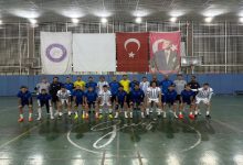 Kapalı bir spor salonunda, Türkiye İşitme Engelliler Futsal Milli Takımı toplu halde kamera karşısında dizilmiş. Mavi ve beyaz formalarla iki sıra halinde poz veriyorlar. Arkada Türk bayrağı ve Atatürk posteri asılı; salon ışıkları parlak ve düzenli.