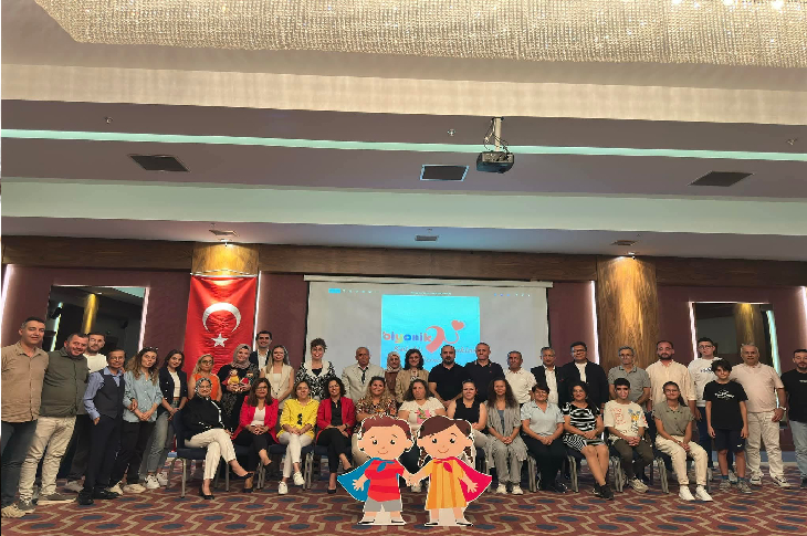 Bir konferans salonunda, sahnede toplu bir hatıra fotoğrafı çekilmiş. Arkada Türk bayrağı ve projeksiyon perdesi yer alıyor. Çoğunluğu yetişkinlerden oluşan kalabalık bir grup yan yana poz verirken, ön tarafta sandalyelerde oturanlar da var. Görselin alt kısmına iki çizgi çocuk figürü eklenmiş.