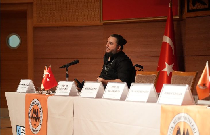 Bir konferans salonunda, önünde mikrofon bulunan bir masada oturan bir konuşmacı görülüyor. Masanın üzerinde isimlikler ve küçük Türk bayrakları yer alırken, arka planda büyük bir Türk bayrağı asılı; ortam resmi bir panel ya da toplantı havası taşıyor.