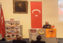 Bir konferans salonunda kürsüde konuşma yapan bir erkek görülüyor. Kürsünün önünde çeşitli dernek logolarının yer aldığı flamalar asılı. Arka duvarda Türk bayrağı ve Mustafa Kemal Atatürk’ün portresi bulunuyor. Sol tarafta bir derneğe ait tanıtım afişi yer alırken, dinleyiciler ön sırada oturmuş şekilde konuşmayı takip ediyor. Ortam resmi bir panel ya da anma programı izlenimi veriyor.