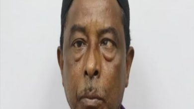 Yakın plan çekilmiş bir erkek portresi görülüyor. Kişi doğrudan kameraya bakıyor. Koyu renk bir bere takıyor ve mor tonlarında bir üst giysi giymiş. Yüz ifadesi ciddi ve nötr; arka plan ise düz, açık renkli ve sade.