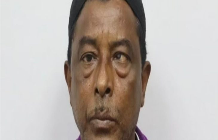 Yakın plan çekilmiş bir erkek portresi görülüyor. Kişi doğrudan kameraya bakıyor. Koyu renk bir bere takıyor ve mor tonlarında bir üst giysi giymiş. Yüz ifadesi ciddi ve nötr; arka plan ise düz, açık renkli ve sade.