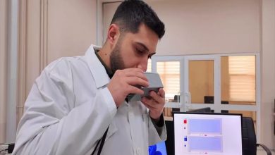 Bir laboratuvar ortamında beyaz önlüklü bir araştırmacı, elindeki küçük cihazı ağzına yaklaştırarak nefes üflüyor. Yan tarafta bilgisayar ekranında ölçüm sonuçlarını gösteren grafikler yer alıyor. Ortam akademik bir çalışma ve test sürecini yansıtıyor.