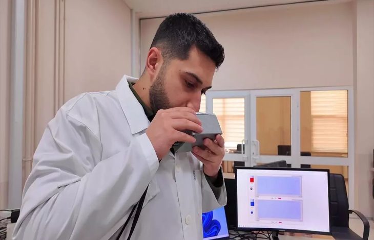 Bir laboratuvar ortamında beyaz önlüklü bir araştırmacı, elindeki küçük cihazı ağzına yaklaştırarak nefes üflüyor. Yan tarafta bilgisayar ekranında ölçüm sonuçlarını gösteren grafikler yer alıyor. Ortam akademik bir çalışma ve test sürecini yansıtıyor.