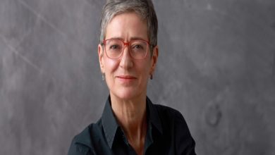 Gri tonlu bir arka plan önünde, kısa gri saçlı, kırmızı çerçeveli gözlük takan bir kişi görülüyor. Koyu renk gömlek giymiş, kameraya sakin ve hafif bir gülümsemeyle bakıyor; portre sade ve resmi bir atmosfer taşıyor.