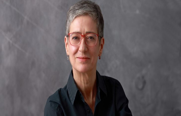 Gri tonlu bir arka plan önünde, kısa gri saçlı, kırmızı çerçeveli gözlük takan bir kişi görülüyor. Koyu renk gömlek giymiş, kameraya sakin ve hafif bir gülümsemeyle bakıyor; portre sade ve resmi bir atmosfer taşıyor.