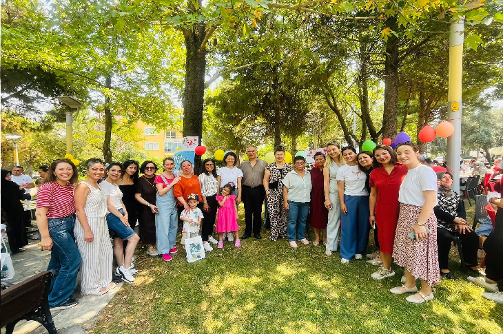 Ağaçların gölgesinde, çimlerin üzerinde kalabalık bir grup birlikte poz veriyor. Çoğunluğu kadınlardan oluşan grup arasında birkaç çocuk da var. Arkada renkli balonlar ve piknik masaları görülüyor; ortam bir açık hava etkinliği ya da kutlama havası taşıyor. Güneşli bir gün, samimi ve neşeli bir atmosfer.