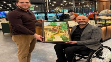 Bir pastane/kafe ortamında iki erkek yan yana poz veriyor. Ayakta duran kişi, tekerlekli sandalyede oturan kişiye çerçeveli bir tablo takdim ediyor. Tabloda taş evler ve çiçekli bir sokak resmi yer alıyor; arka planda tatlı tezgâhları ve çalışanlar görülüyor.