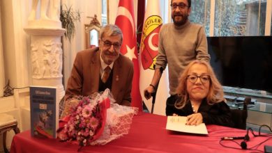 Bir salonda, önünde masa bulunan bir imza günü ya da tanıtım etkinliği görülüyor. Masada çiçek buketi ve kitap yer alıyor; iki kişi oturuyor, arkalarında ayakta duran bir kişi bulunuyor. Arka planda Türk bayrağı ve bir kuruma ait bayrak yer alıyor; ortam resmi ve saygın bir buluşmayı yansıtıyor.