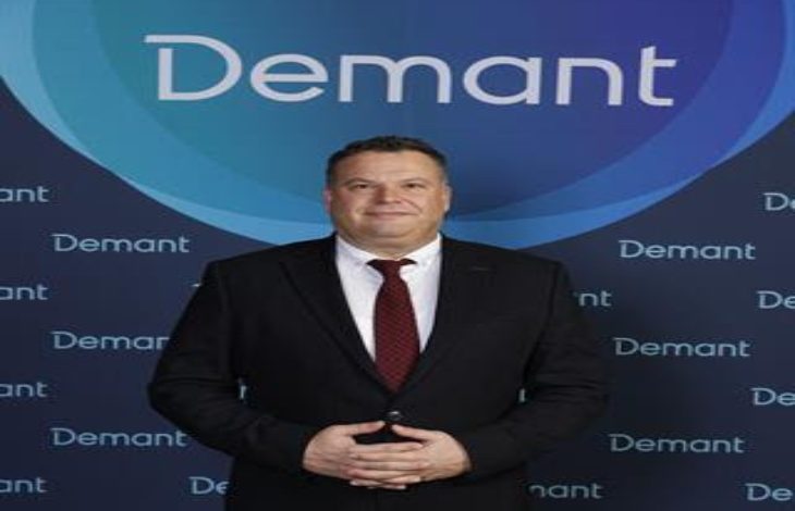 Koyu renk takım elbise ve bordo kravat takan bir erkek, üzerinde “Demant” logosu bulunan mavi tonlardaki bir fon önünde ayakta duruyor. Ellerini önde birleştirmiş, kameraya doğru sakin ve resmi bir poz veriyor.