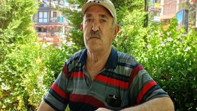 Açık havada, yeşillikler arasında oturan yaşlı bir adam görülüyor. Üzerinde çizgili tişört ve şapka var; sakin ama yorgun bir ifadeyle kameraya bakıyor. Arka planda ağaçlar ve şehir yapıları seçiliyor.