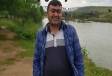 Bir nehir kıyısında açık havada çekilmiş bir fotoğraf. Önde duran bir erkek kameraya gülümseyerek poz veriyor. Üzerinde mavi mont ve polo yaka bir tişört var. Arka planda su kenarı, yeşillik alan ve bir köprü görünüyor.