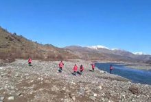 Taşlık bir nehir kıyısında, kırmızı yelekli kişiler dağlık bir bölgede arama yaparak ilerliyor. Arka planda karla kaplı zirveler görülüyor.