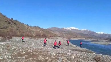 Taşlık bir nehir kıyısında, kırmızı yelekli kişiler dağlık bir bölgede arama yaparak ilerliyor. Arka planda karla kaplı zirveler görülüyor.