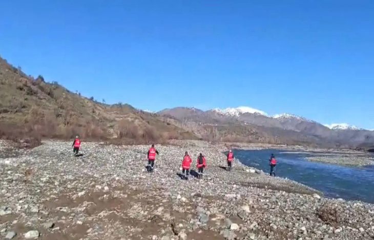 Taşlık bir nehir kıyısında, kırmızı yelekli kişiler dağlık bir bölgede arama yaparak ilerliyor. Arka planda karla kaplı zirveler görülüyor.