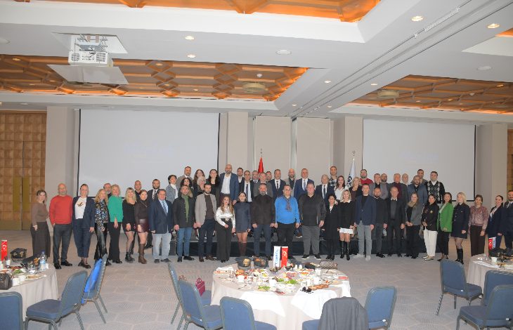 Geniş bir otel salonunda, ön tarafta yuvarlak masalar ve sandalyeler, arkada ise sahne önünde yan yana dizilmiş kalabalık bir grup görülüyor. Grup, resmi bir etkinlik kapsamında toplu fotoğraf için poz veriyor; salonun tavanı geometrik desenli ve aydınlatmalarla donatılmış.