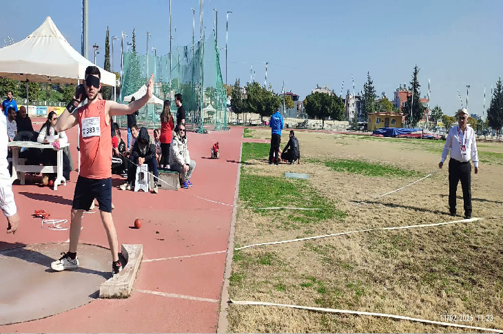Atletizm sahasında bir sporcu gülle atma alanında atışa hazırlanıyor. Kolunu geriye almış, denge için diğer kolunu açmış durumda. Yan tarafta hakem ölçüm için bekliyor; arka planda izleyiciler ve saha ekipmanları görülüyor. Yarış anının tam eşiği.