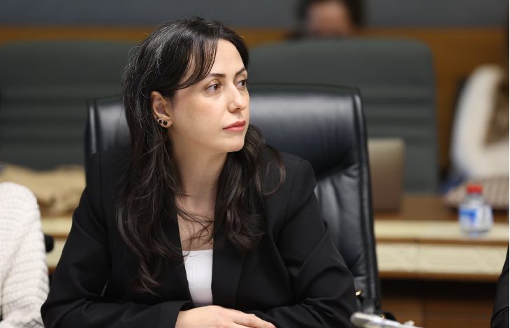 Bir toplantı salonunda oturan bir kadın, ciddi ve dikkatli bir ifadeyle konuşmaları dinlerken görülüyor.